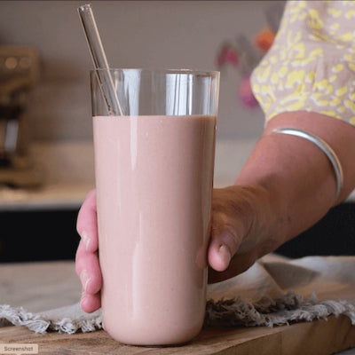 Hand holding a glass of pink smoothie with a straw on a wooden surface.