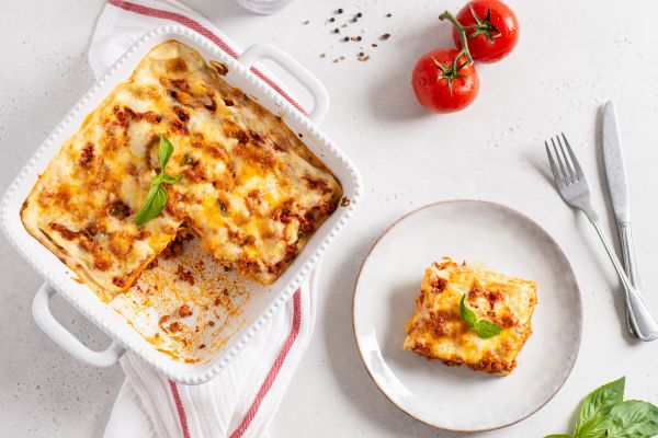 Baked lasagna in a dish with a serving on a plate, surrounded by tomatoes and basil.