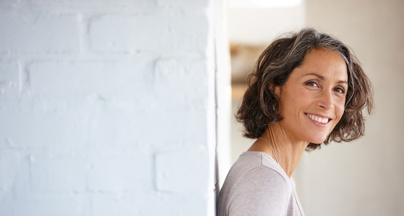 A smiling woman with short brown hair looks over her shoulder from behind a doorway, wearing a light gray top against a white brick wall.