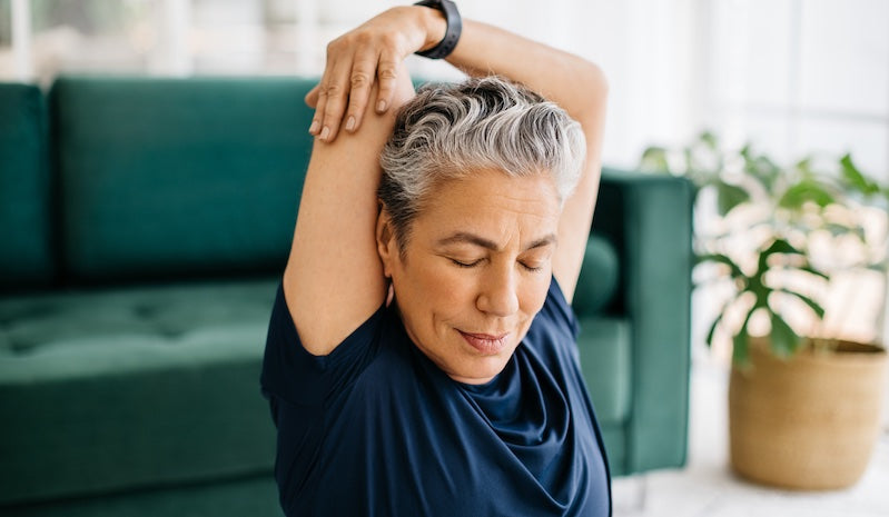 A woman with short gray hair wearing a navy blue t-shirt stretches her arm over her head in a bright living room with a green couch and a potted plant.