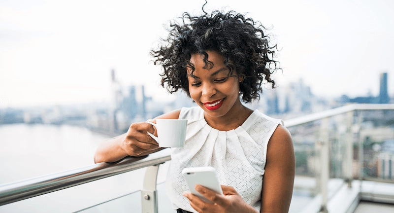 A smiling woman with curly black hair stands on a balcony overlooking a city skyline, holding a white mug in one hand and a white smartphone in the other.