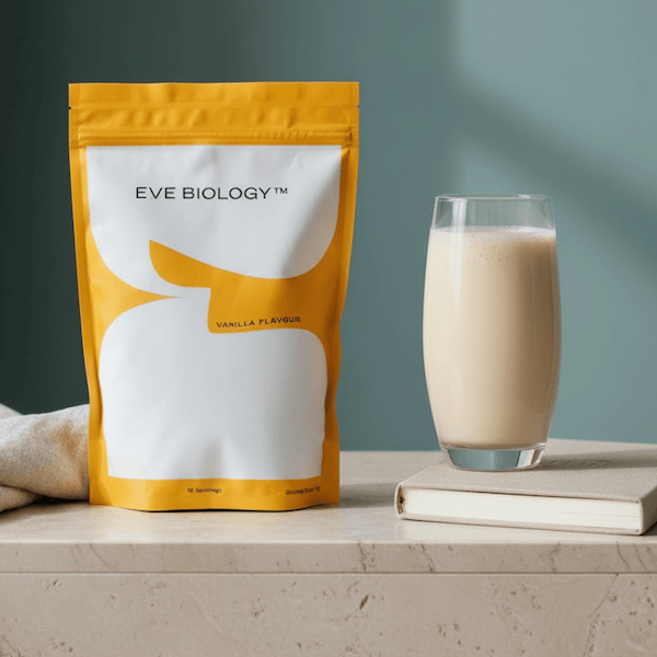 A stand-up pouch of Eve Biology Vanilla Flavour powder sits on a light stone surface next to a tall glass of creamy beige drink and a small book, against a muted blue-green wall.