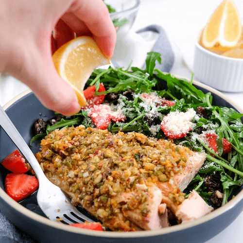 Salad with salmon, strawberries, and arugula being garnished with lemon.