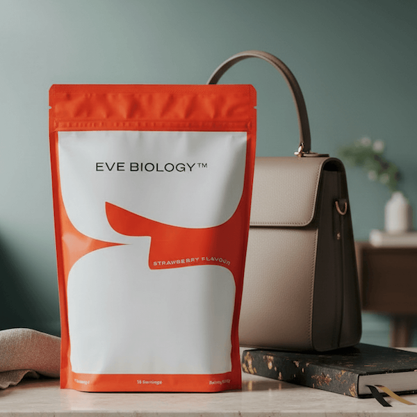 A stand-up pouch of EVE BIOLOGY strawberry-flavour product stands on a tabletop next to a woman's bag. The pouch is bright orange and white with black branding text.