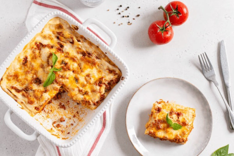Baked lasagna in a dish with a serving on a plate, accompanied by tomatoes and utensils.
