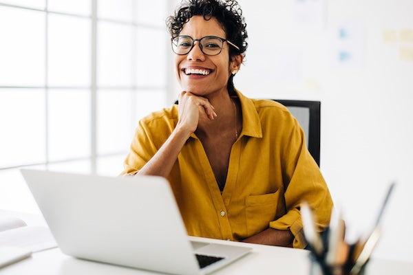 Woman in yellow shirt sitting at a desk with a laptop, smiling.