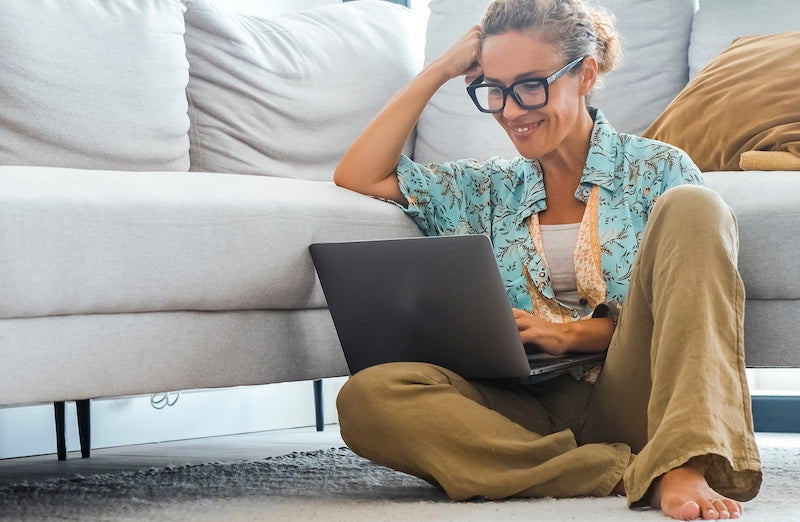 A woman with glasses sits on a gray sofa floor cushion, smiling and using a laptop in a bright living room.