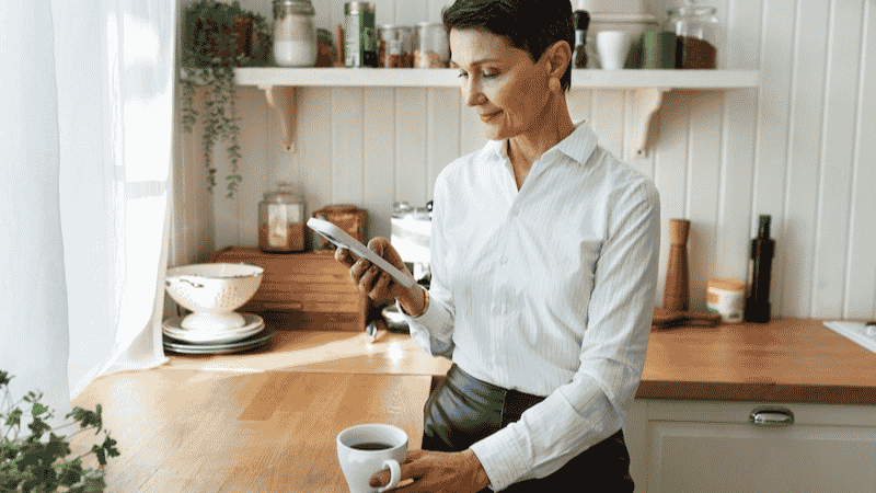 Woman in her kitchen drinking coffee reading her phone.
