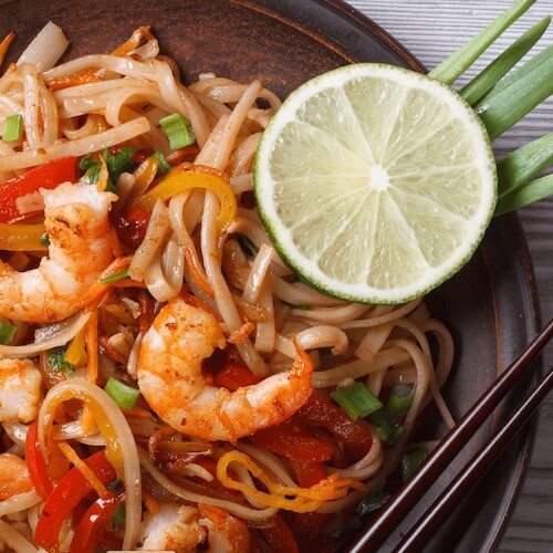 Noodle dish with shrimp, vegetables, and a lime wedge on a wooden plate.