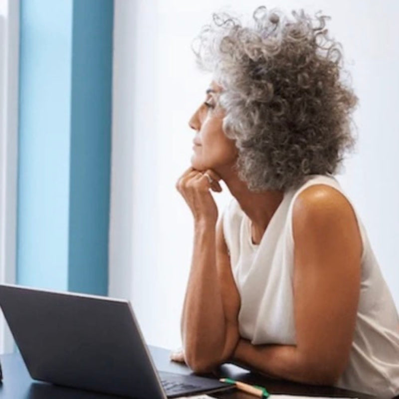 Woman with gray hair sitting at a desk with a laptop, looking thoughtful.