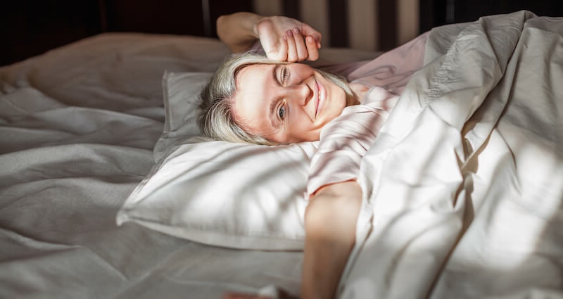 A smiling older woman lies in bed under soft white and beige bedding, with sunlight casting gentle shadows across her face and the duvet.