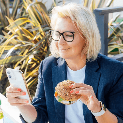 Woman holding a sandwich and phone outdoors with plants in the background