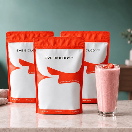 Three red and white stand-up pouches of Eve Biology supplement arranged on a marble countertop, with a pink strawberry smoothie in a clear glass beside them against a teal wall.
