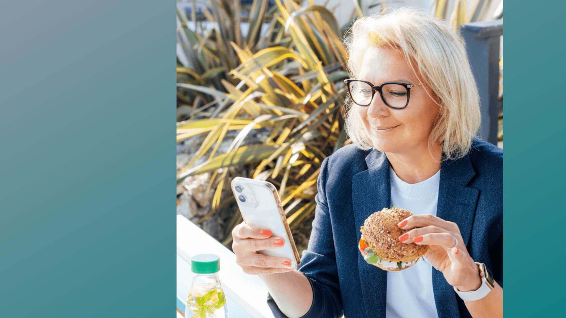 Woman holding a smartphone and a sandwich outdoors with plants in the background