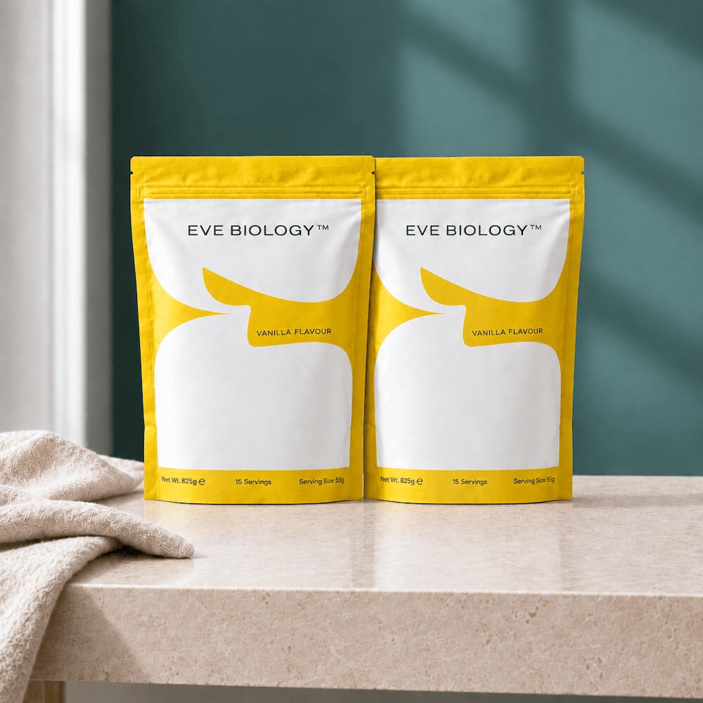 Two yellow and white stand-up pouches of Eve Biology Vanilla Flavour powder sit side-by-side on a light beige marble countertop with a beige towel draped on the left, against a dark teal wall.