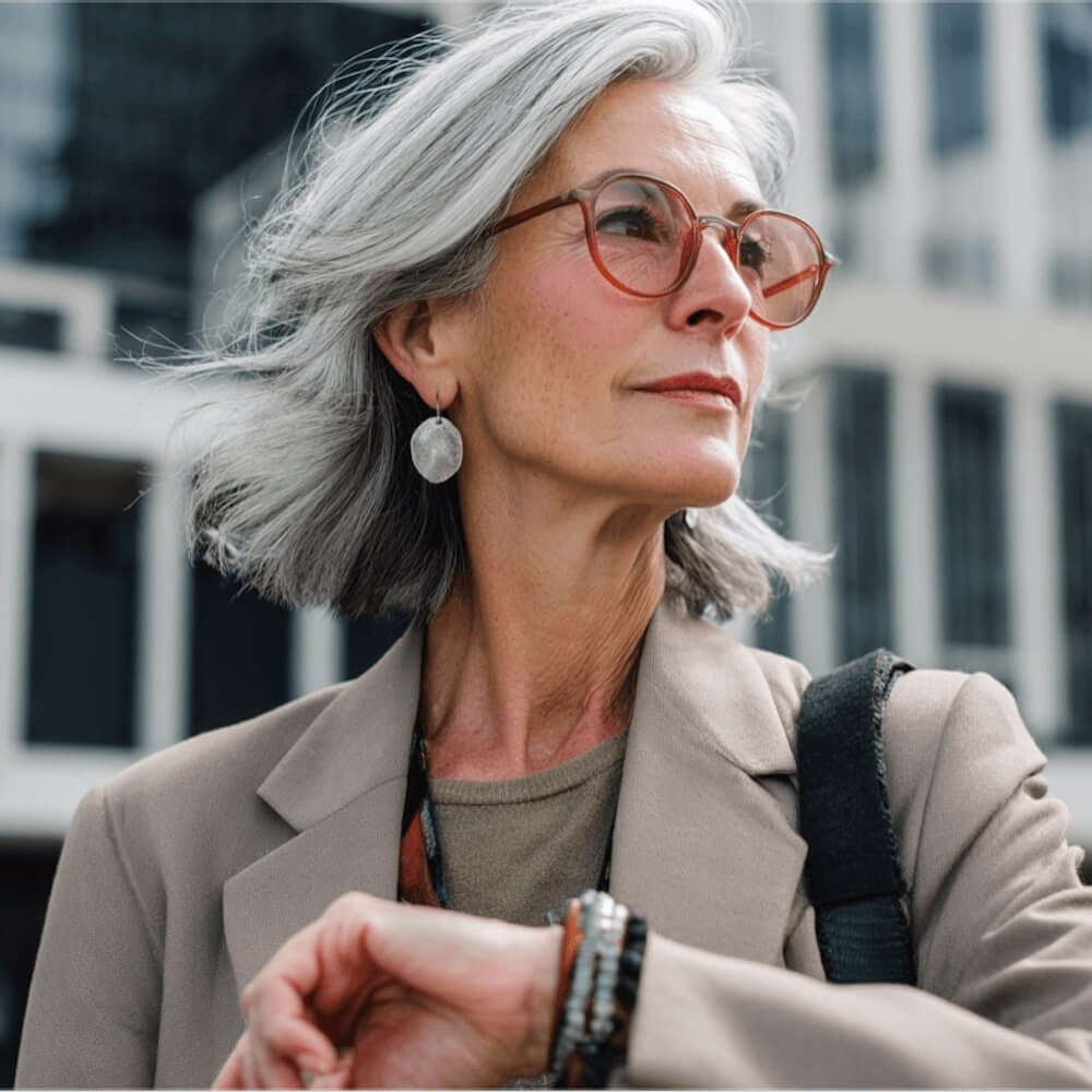 Woman with gray hair and glasses looking to the side, wearing a beige jacket and a black strap over her shoulder.
